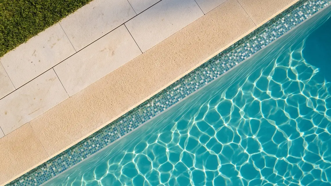 Vista aérea del borde de una piscina cruzando el encuadre en diagonal: el agua con sus cáusticas, una franja de mosaico bajo la línea de agua, el borde de árido granulado y una terraza de pastelones claros