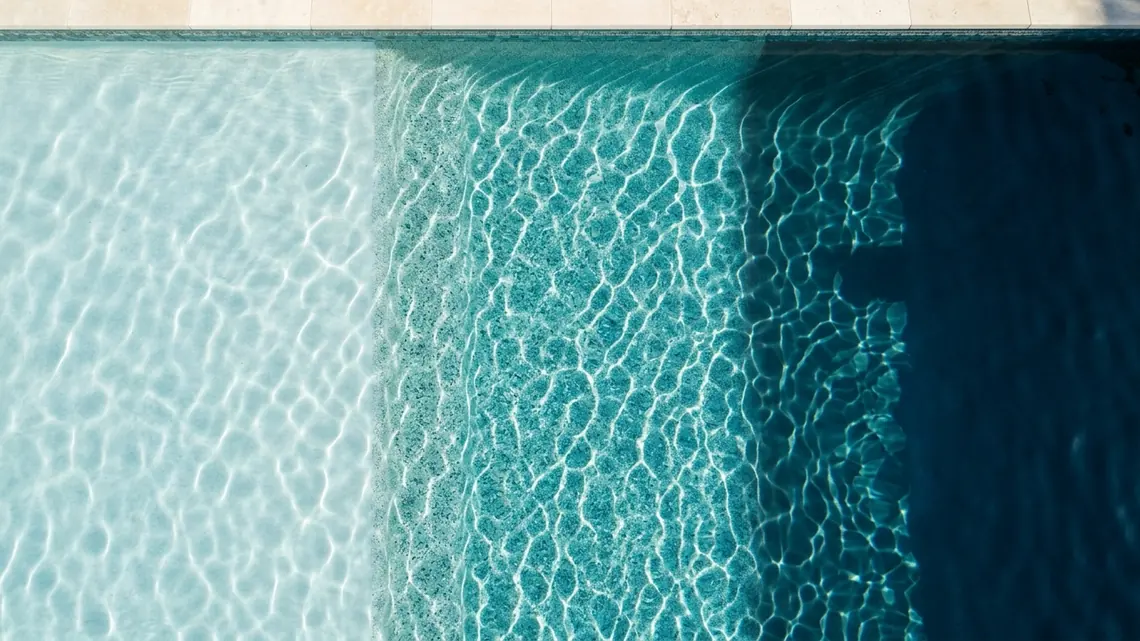 Fondo de piscina visto desde arriba a través del agua, donde el revestimiento cambia en tres franjas: un enlucido claro, una superficie de cuarzo granulado y una terminación oscura y lisa